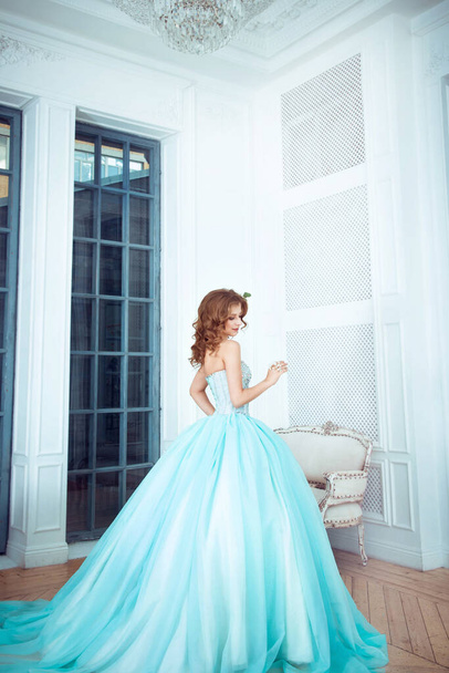 The beautiful young girl the princess in a long magnificent turquoise dress stands in a light castle. Fairytale fashion photo session Cinderella. Beautiful room.  - Фото, зображення