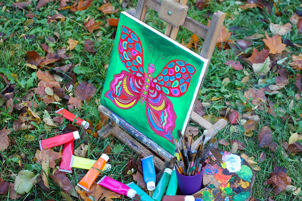 Easel com tintas e pintura com borboleta sobre tela no parque de outono
. - Foto, Imagem