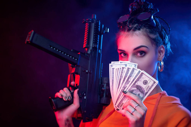 beautiful face brunette in a red jumpsuit with a short-barrelled machine gun on a dark background in neon light. keeps money dollars - Valokuva, kuva