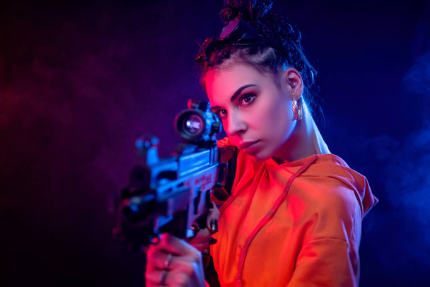 beautiful face brunette in a red jumpsuit with a short barrelled machine gun on a dark background in neon light - Photo, Image