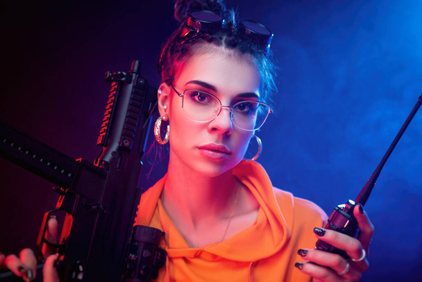 heldere brunette in een oranje jumpsuit met een machinegeweer op een donkere achtergrond in neon licht. praten op de radio - Foto, afbeelding