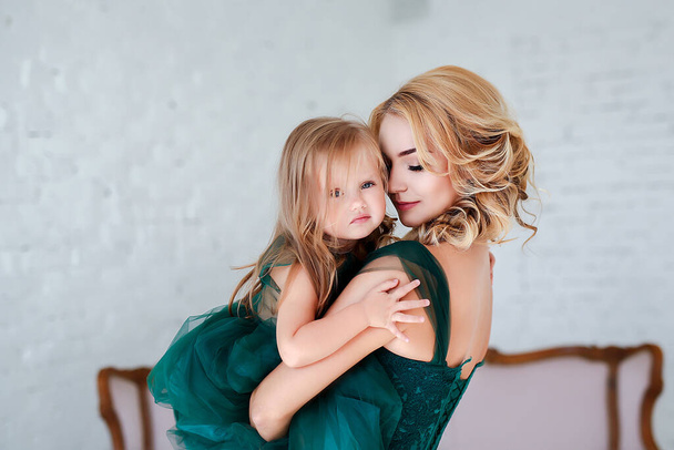 Portret pięknej młodej matki z uroczą blondynką siedzącą w białym wnętrzu ubraną w eleganckie zielone sukienki. - Zdjęcie, obraz