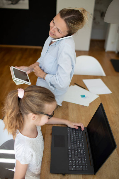 woman working from home office and talking by phone with her daughter homeschooling with laptop during corona crisis - Photo, Image