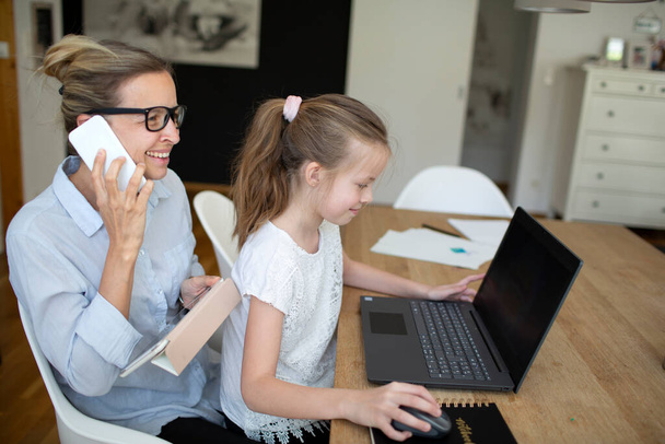 woman working from home office and talking by phone with her daughter homeschooling with laptop during corona crisis - Zdjęcie, obraz
