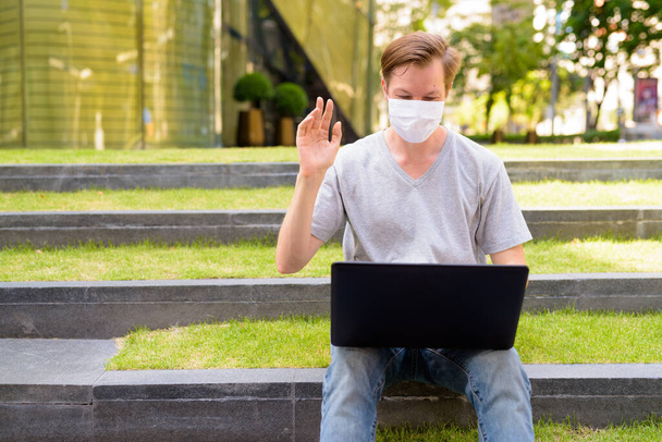 Portrait of young Scandinavian man with mask for protection from corona virus outbreak outside modern building with nature - Fotografie, Obrázek