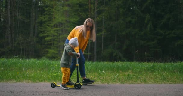 Junge Mutter zeigt ihrem kleinen Sohn, wie man mit dem Roller im Park fährt. Aktive Familienfreizeit. Training, sportliche Fähigkeiten, die ersten Schritte im Sport. Zeitlupe - Filmmaterial, Video