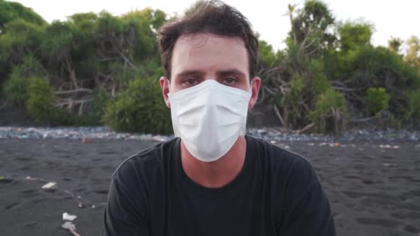 Mladý muž sedí na pláži na Bali, nosí ochranu obličeje v prevenci koronaviru. Obsah: Uzamčení v Indonésii, Žádné cestování, Těžké časy, Čekání na lepší budoucnost. - Záběry, video