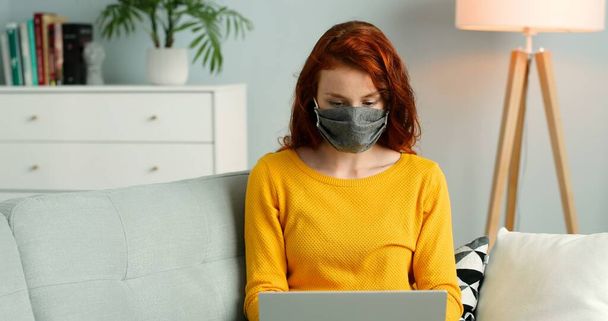 Cheerful young redhead girl in mask sitting on sofa in living room at home and using silver laptop. - Фото, изображение