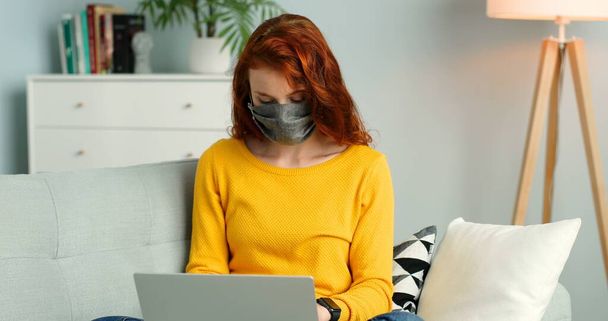 Cheerful young redhead girl in mask sitting on sofa in living room at home and using silver laptop. - Φωτογραφία, εικόνα