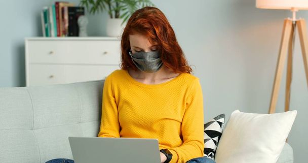 Cheerful young redhead girl in mask sitting on sofa in living room at home and using silver laptop. - Fotó, kép