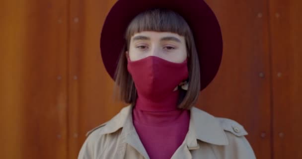 Close up shot of good looking girl in protective cotton mask turning head and looking to camera. Portrait of millennial woman in hat with reusable mask on face . Zoom in. Rust background. - Кадри, відео