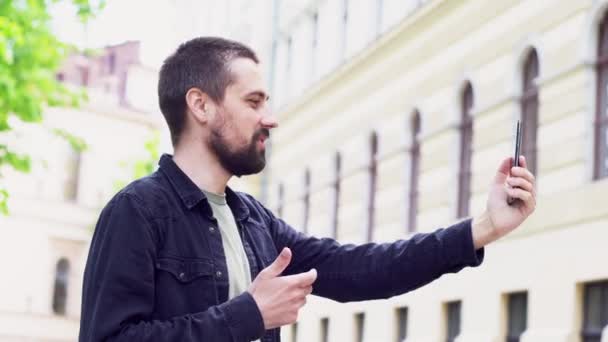 Een jonge blanke man met een baard houdt een telefoon op zijn uitgestrekte hand en praat. De blogger communiceert per videogesprek. De man glimlacht op straat. - Video
