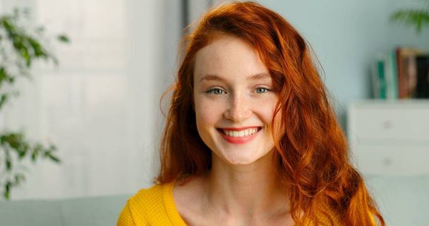 Portrait of beautiful cheerful redhead girl smiling looking at camera sitting on sofa at home. - Foto, Imagem