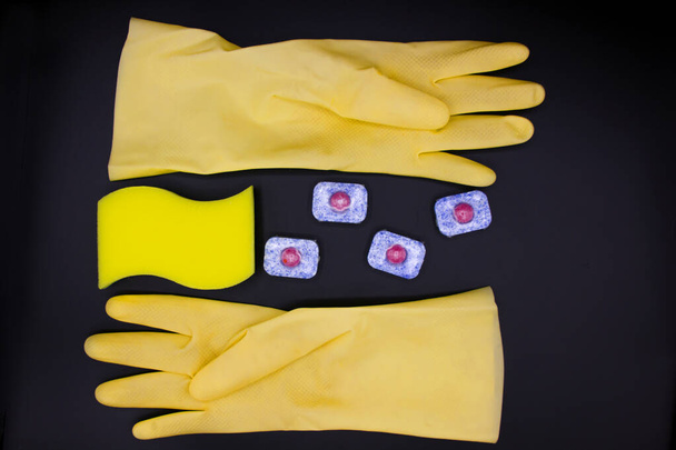 Geschirrspülmittel: Schwamm, Geschirrspültablette, gelbe Handschuhe auf schwarzem Hintergrund. Von oben gesehen, aus nächster Nähe. Sauberes Haus und Pflegekonzept - Foto, Bild