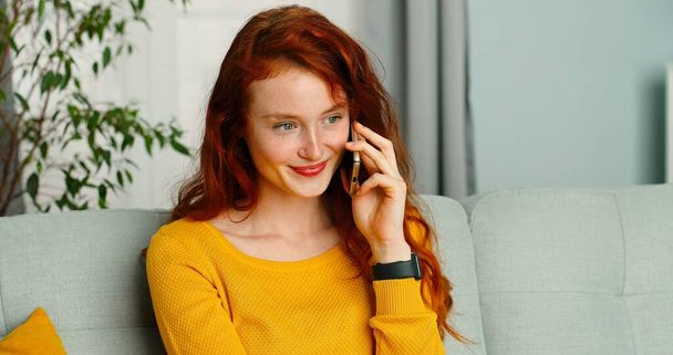 Happy and positive girl sitting on sofa at home and talking on her smartphone. Communication app smartphone concept - Фото, изображение
