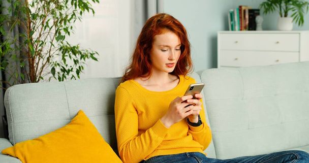 Beautiful young red-haired woman with curly hair texting message on smartphone at home. - Photo, image