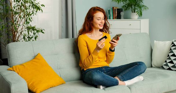 Jonge mooie roodharige vrouw zittend op de bank in de woonkamer en online winkelen op mobiele telefoon. - Foto, afbeelding