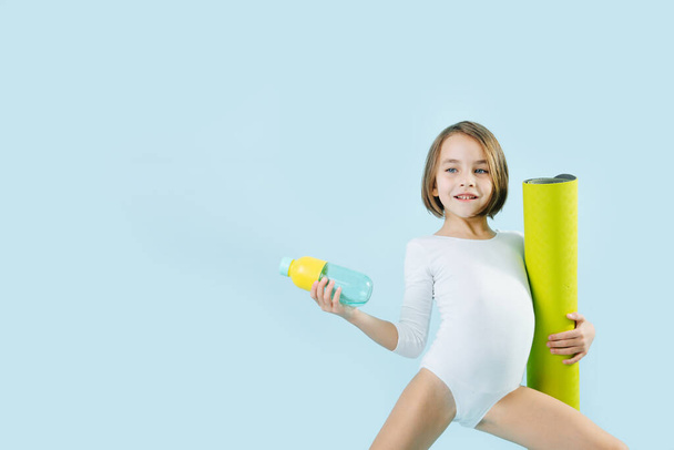 Little girl in a white leotard with rolled mat and water bottle over blue background. Standing in a gymnastic position. - Foto, afbeelding