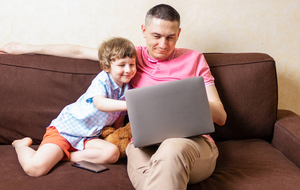 Szczęśliwy młody ojciec siedzieć na komputerze za pomocą laptopa zrelaksować się z synem przedszkolak baw się dobrze razem, uśmiechnięty tata i mały chłopiec dziecko cieszyć weekend w domu odpocząć na kanapie zajęty gadżetami. Edukacja online. - Zdjęcie, obraz