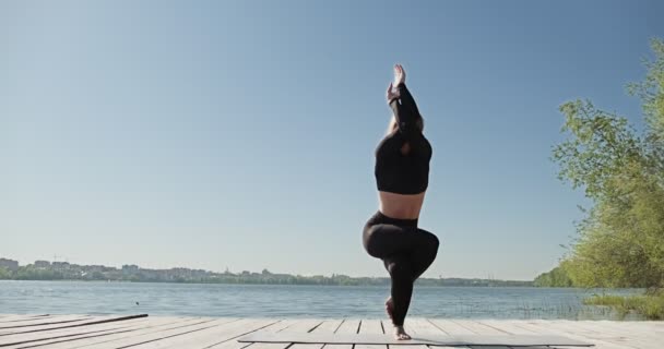 Egy fiatal szőke nő jógázik a fapadlón a tónál. Egyszeri sport egészséges képzés a természetről napsütéses időben - Felvétel, videó