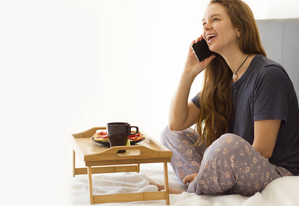 Veselá žena snídá v posteli. Čaj a palačinky s jahodou. Asijská běloška se usmívá, mluví na mobilu, dívá se nahoru a směje - Fotografie, Obrázek