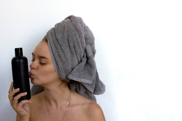 Jovem mulher em toalha cinza na cabeça sobre o fundo branco detém e beijar garrafa de xampu preto sem logotipo. Mulher em banho com pele limpa e macia
. - Foto, Imagem