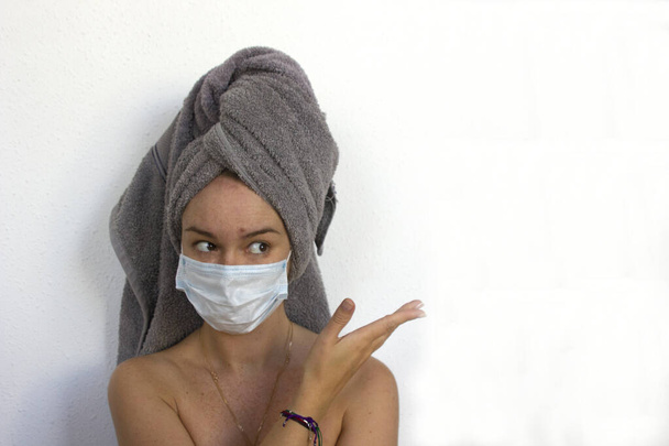 jonge vrouwen met een blauw medisch masker en een grijze handdoek op haar hoofd tonen iets, ruimte voor tekst - Foto, afbeelding