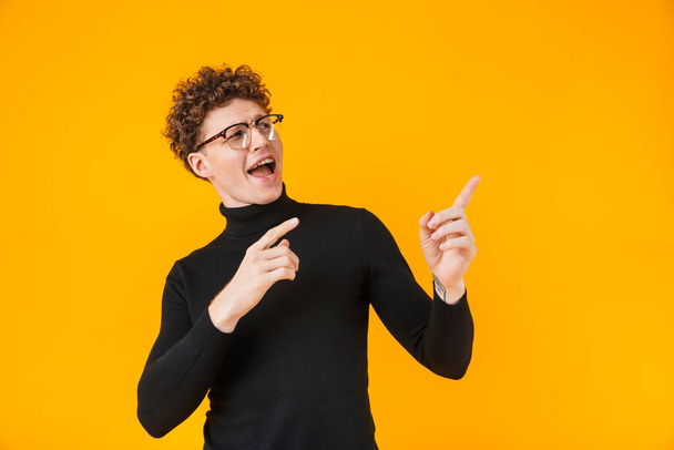 Image of young excited man in eyeglasses pointing fingers aside isolated over yellow background - Foto, Bild