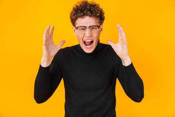 Image of young angry man in eyeglasses screaming at camera with throwing up hands isolated over yellow background - Foto, afbeelding