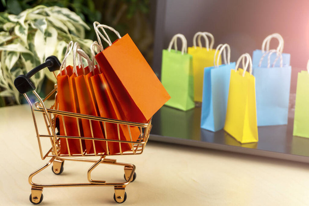 online vásárlási koncepció. kis bevásárlókocsi színes bevásárlószatyrokkal belsejében stand laptop közelében, kis kézbesítő dobozok, bankkártya - Fotó, kép