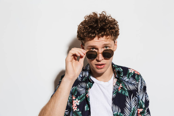 Image of young confused man holding sunglasses and looking at camera isolated over white background - Foto, imagen