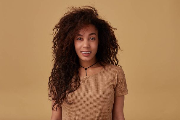 Indoor shot of pleased young curly dark skinned brunette lady dressed in nude t-shirt looking at camera with charming smile while posing over beige background - Foto, immagini