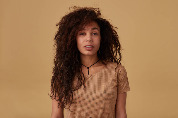 Portrait of young long haired curly dark skinned female with voluminous hairstyle looking calmly at camera while standing over beige background in casual clothes - Photo, Image