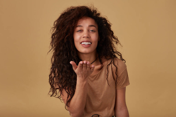 Cheerful young pretty long haired curly female with dark skin keeping her palm raised while sending air kiss to camera and smiling gladly, isolated over beige background - Фото, изображение