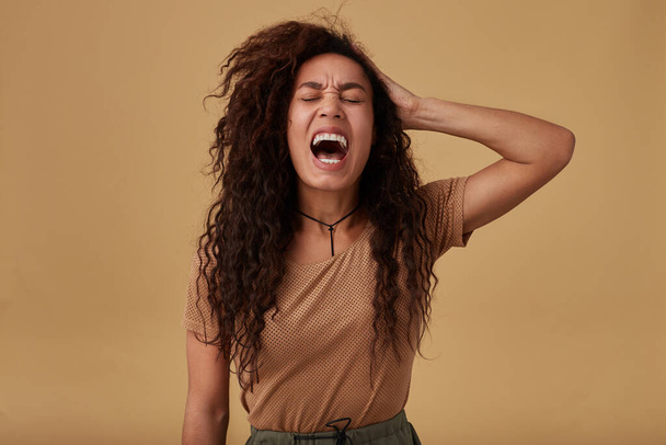 Pissed-off young long haired curly dark skinned female keeping her eyes closed while screaming emotionally and holding hand on her head, isolated over beige background - Φωτογραφία, εικόνα