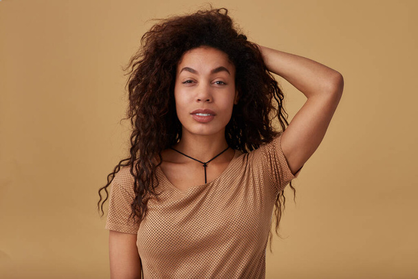 Portrait of charming young long haired curly dark skinned female with voluminous hairstyle raising hand to her head and looking calmly at camera, standing over beige background - Foto, imagen