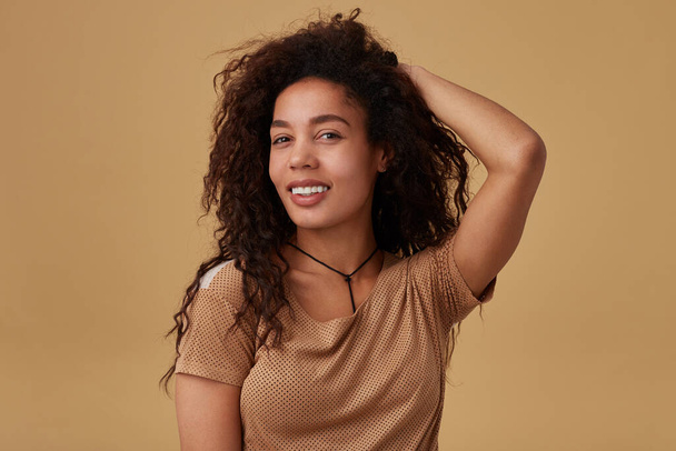 Portrait of young glad long haired curly dark skinned lady looking positively at camera with charming smile while keeping raised hand on her head, isolated over beige background - Photo, Image