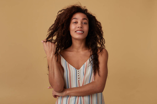 Glad young attractive long haired curly dark skinned woman twisting her hair on finger while looking positively at camera, posing over beige background - Photo, Image