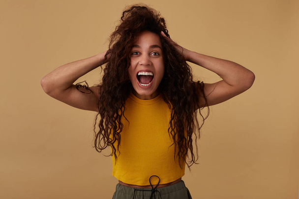 Expressive young pretty brown haired curly brunette lady with dark skin keeping her mouth wide opened while clutching her head with raised hands, posing over beige background - Photo, Image