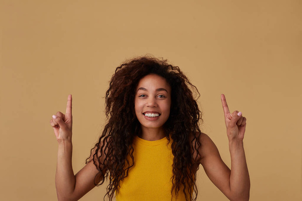Studio laukaus nuorten iloinen ruskea tukka kihara tumma iho nainen rento kampauksen hymyillen laajasti samalla osoittaa ylöspäin etusormet, eristetty yli beige tausta
 - Valokuva, kuva