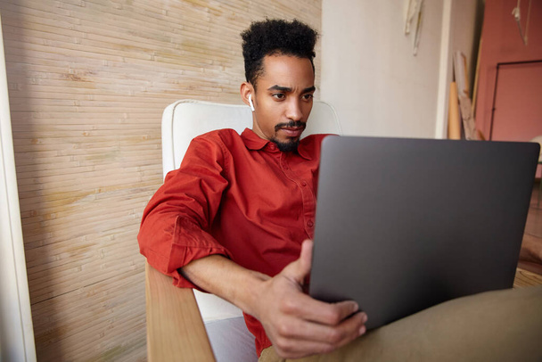 Indoor photo of young bearded brunette male with dark skin looking attentively at screen of his notebook while working remotely from home, isolated over beige interior - Photo, image