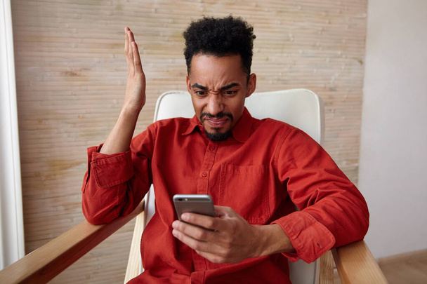 Confused young bearded dark skinned male with short haircut grimacing his face and keeping hand raised while looking on his smartphone with pout, posing over beige background - Fotó, kép