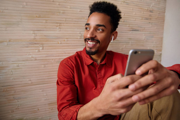 Portrét mladého radostného krátkosrstého vousatého bruneta, který drží v rukou mobilní telefon a usmívá se, zatímco se dívá stranou, sedí v interiéru domu - Fotografie, Obrázek