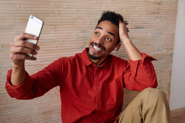 Excited young bearded dark skinned male with beard keeping hand raised while having pleasant phone talk and smiling widely, posing over beige interior - Foto, Imagem