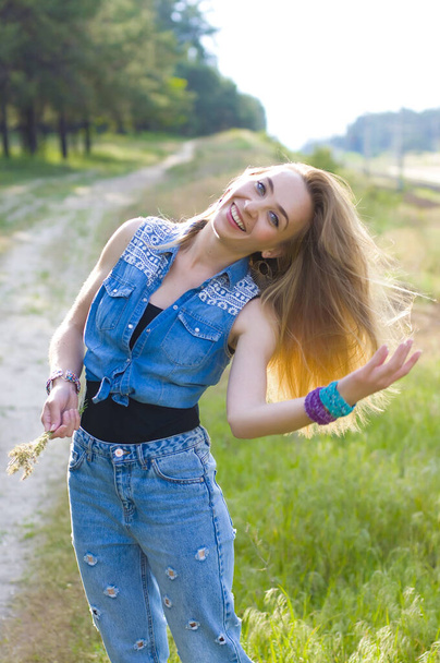 cute blonde in jeans clothes stands on a path on the outskirts of a summer forest with hair fluttering in the wind and smiles at the camera - Photo, Image