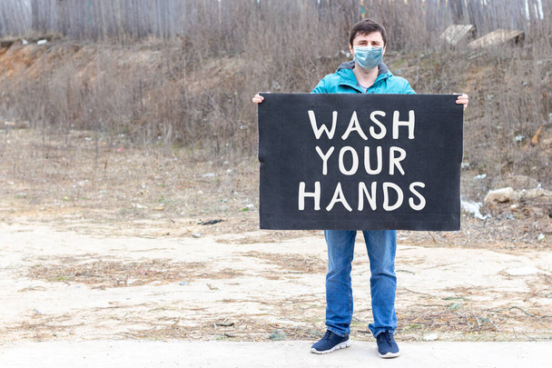 Mężczyzna w masce ochronnej medycznej twarzy z czarnym kawałkiem tkaniny z napisem "umyć ręce" na rozmazanym tle martwej natury ze śmieciami. Koncepcja kontroli zanieczyszczeń i kwarantanny - Zdjęcie, obraz