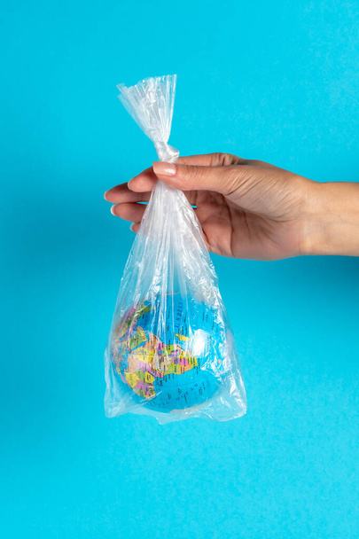 Female hand holding a plastic bag with Earth globe on blue background. Minimal ecology concept. - Фото, зображення