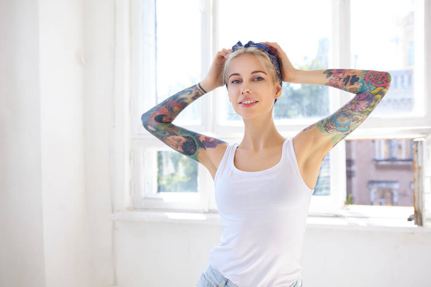 Sunny shot of young attractive tattooed blonde lady keeping raised hands on her head while looking at camera with pleasant smile, standing in front of big window - Valokuva, kuva