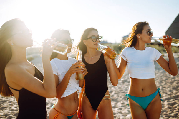 Jonge meisjes in bikini hebben plezier en drinken bier op het strand. Vriendinnen genieten op strandvakantie. Zomer, relax en lifestyle concept. - Foto, afbeelding