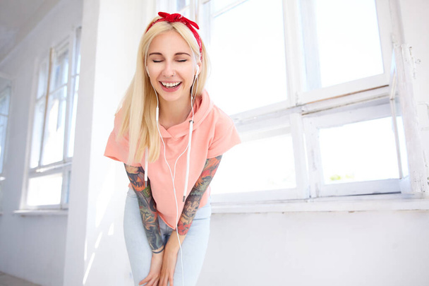 Joyeux jeune belle dame blonde tatouée en bandeau et t-shirt rose gardant les yeux fermés tout en souriant agréablement, posant sur studio lumineux en vêtements décontractés
 - Photo, image